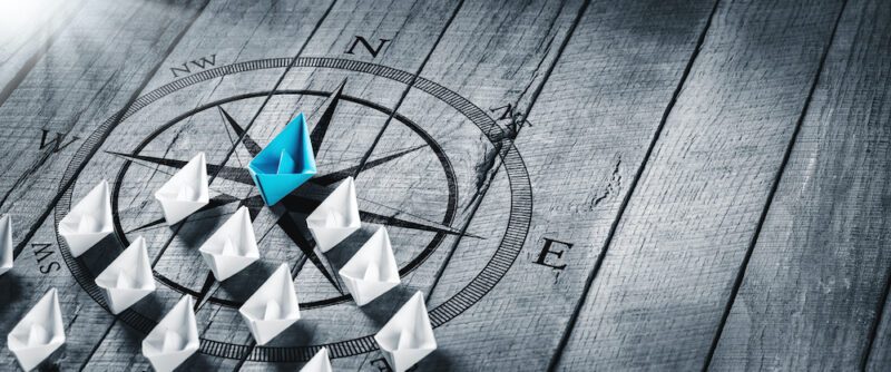 Blue Paper Boat Leading A Fleet Of Small White Boats With Compass Icon On Wooden Table With Sunlight - Leadership Concept